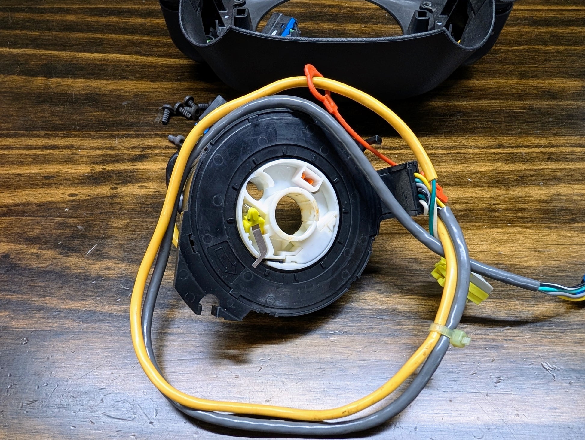 A close-up of a General Motors KIT Steering Wheel Radio Controls & Clock Spring for 1999 Chevy S10, Blazer, and more, with attached wires on a wooden surface alongside screws and a detached plastic part.