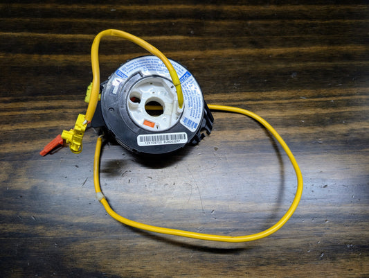 A General Motors OEM Steering Wheel Air Bag SRS Clock Spring, tested for 2004-2005 Chevy S10 Blazer and more, with a yellow wire and connectors, rests on a wooden surface; it has a barcode and multiple warning labels.