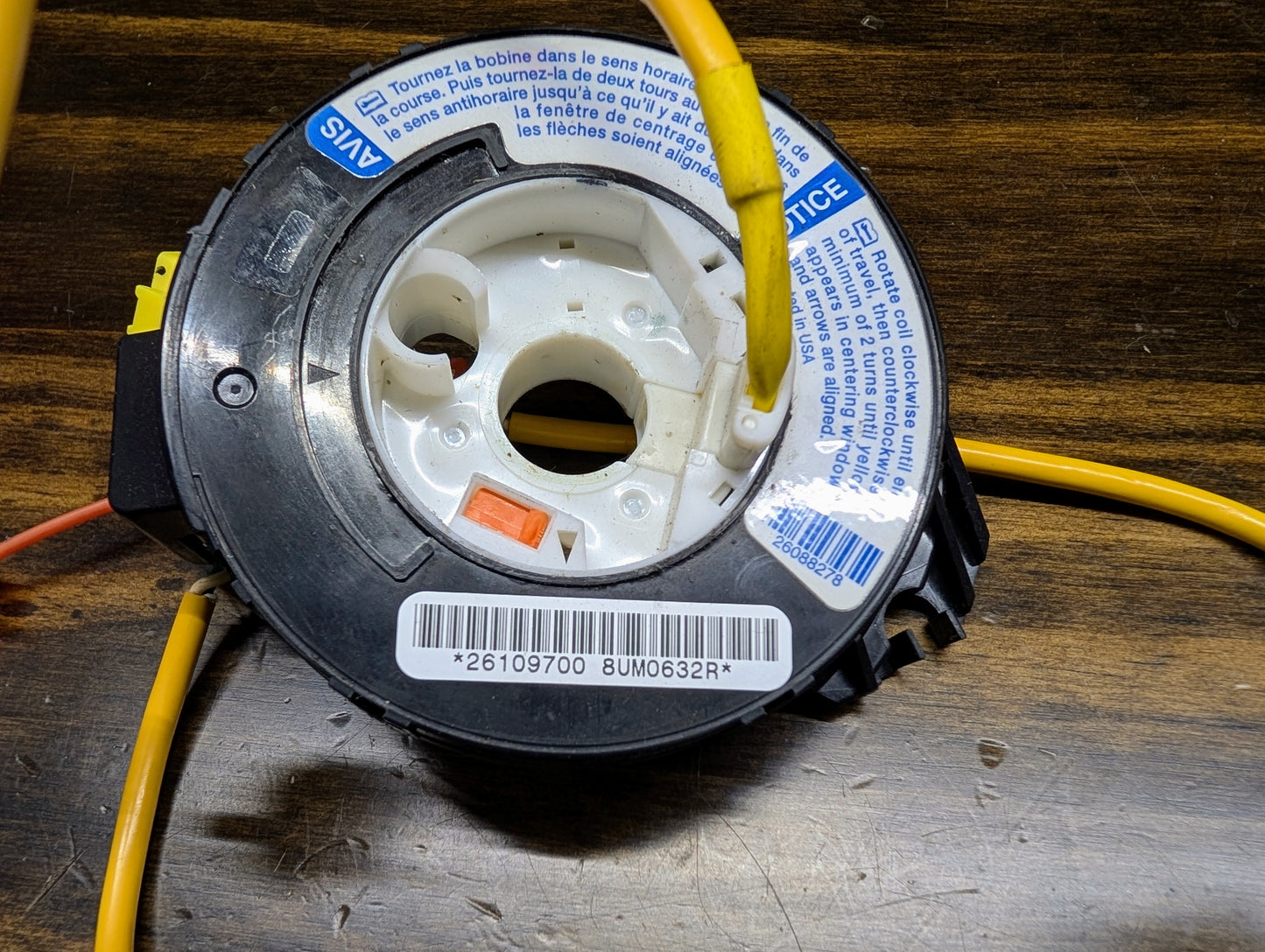 A close-up of the General Motors OEM Steering Wheel Air Bag SRS Clock Spring, tested for 2004-2005 Chevy S10 Blazer and more, with yellow wires, barcode sticker, and bilingual warning labels on a wooden surface.