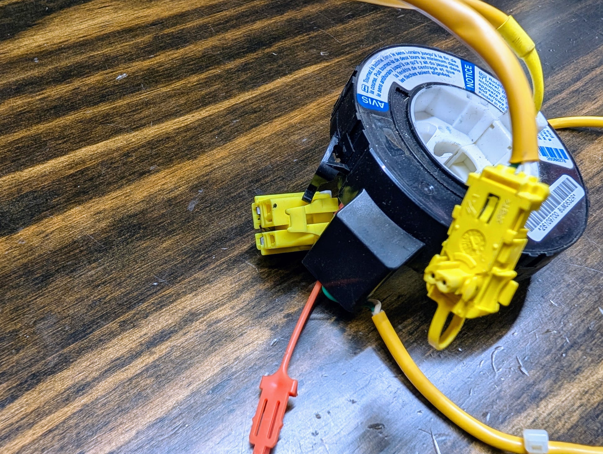 A General Motors OEM Steering Wheel Air Bag SRS Clock Spring for 2004-2005 Chevy S10 Blazer is shown on a wooden surface. It has attached wiring harnesses with yellow and red connectors and a white label with blue text.