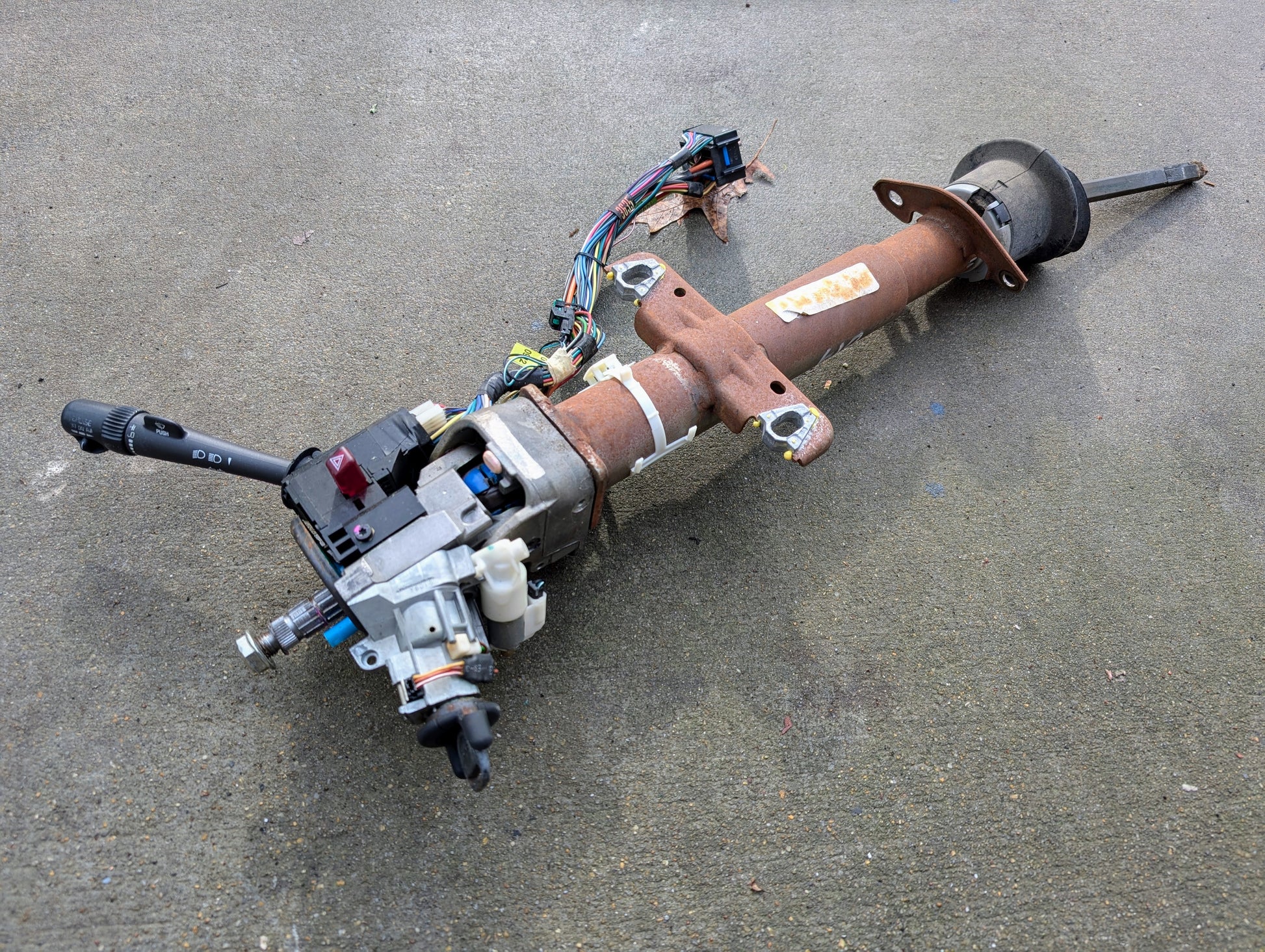 A General Motors OEM Steering Column w/Tilt, Cruise Control, and Console Shift for 1998-2005 Chevy S10/Blazer and more, showing wear and age, with attached wiring and ignition switch, is lying on a concrete surface.