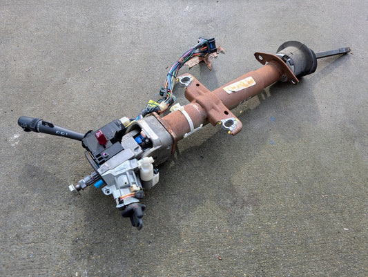 A General Motors OEM Steering Column w/Tilt, Cruise Control, and Console Shift for 1998-2005 Chevy S10/Blazer and more, showing wear and age, with attached wiring and ignition switch, is lying on a concrete surface.