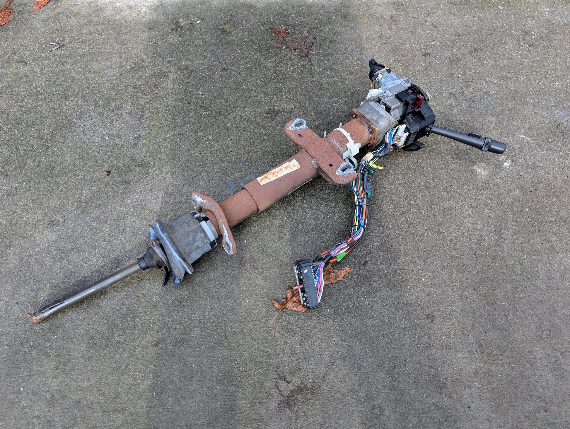 A General Motors OEM steering column with tilt, cruise control, and console shift for 1998-2005 Chevy S10 Blazer and more rests on concrete alongside attached wiring and switches.