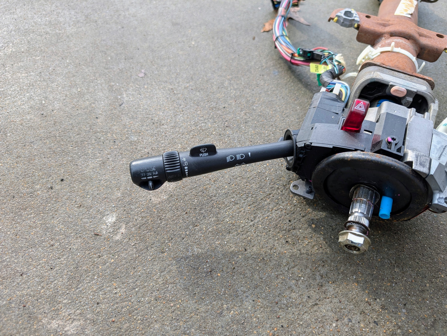 A General Motors OEM Steering Column w/Tilt, Cruise Control, and Console Shift for 1998-2005 Chevy S10 Blazer and more, with attached wiring harnesses, lies on concrete.
