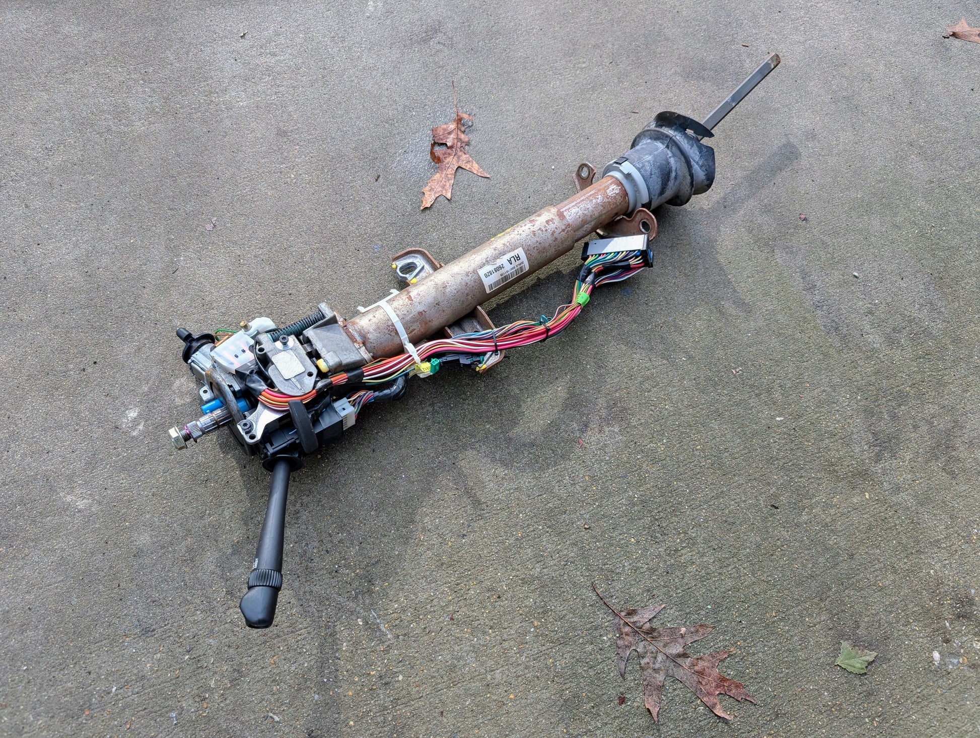 A General Motors OEM Steering Column w/Tilt, Cruise Control, and Console Shift for 1998-2005 Chevy S10 Blazer and more lies outdoors on concrete with exposed wiring, attached controls, and a few brown leaves nearby.
