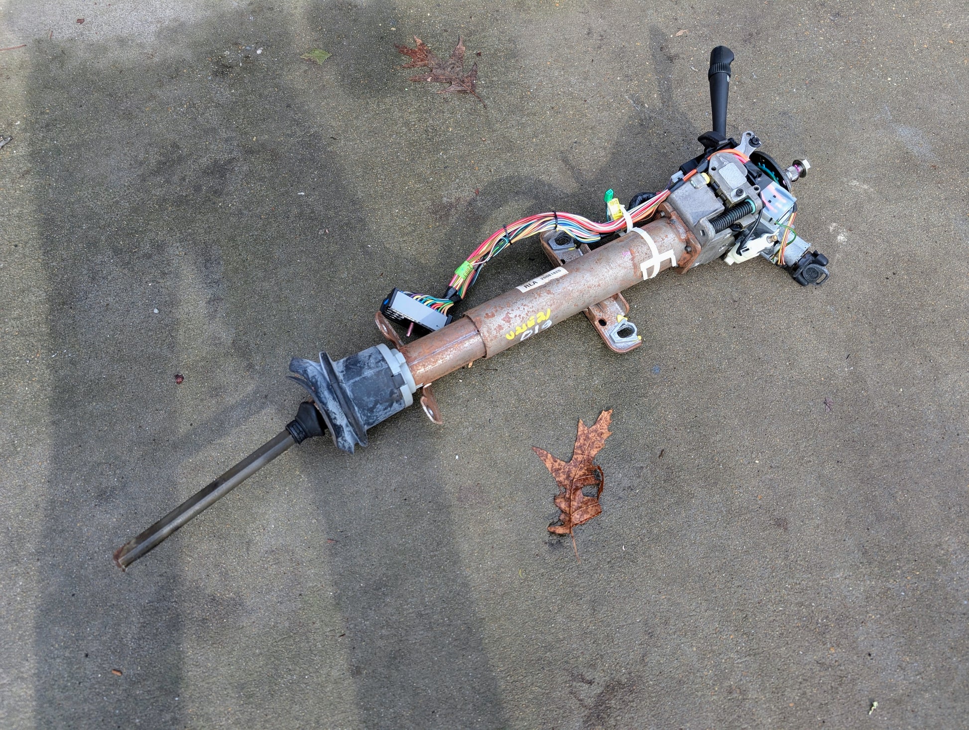 A General Motors OEM Steering Column w/Tilt, Cruise Control, and Console Shift for 1998-2005 Chevy S10 Blazer rests disassembled on concrete next to a dried brown leaf.