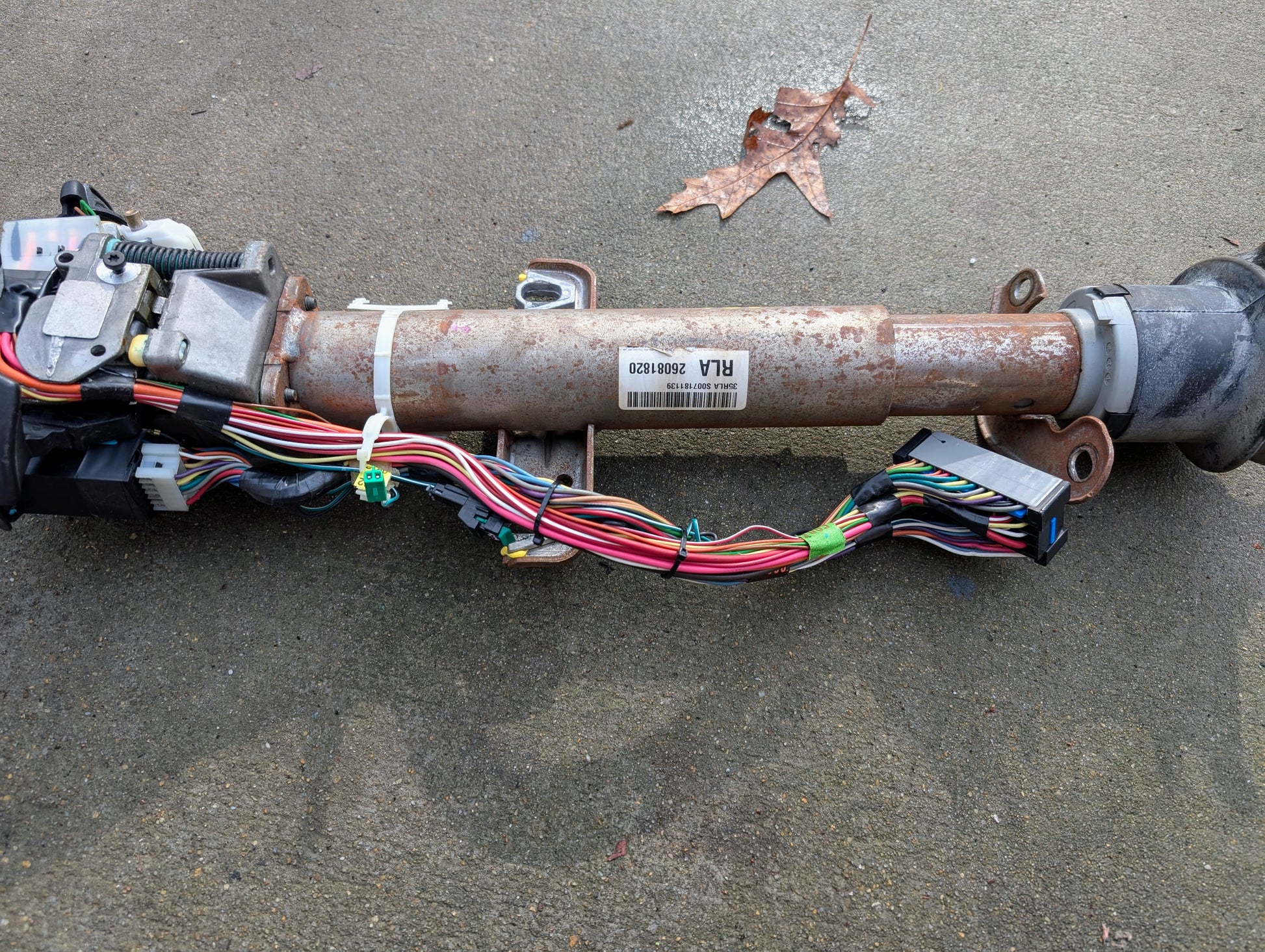 A General Motors OEM steering column with tilt, cruise control, console shift, and wiring for 1998-2005 Chevy S10 Blazer and more rests on concrete with visible rust and barcode sticker; a dried leaf lies nearby.