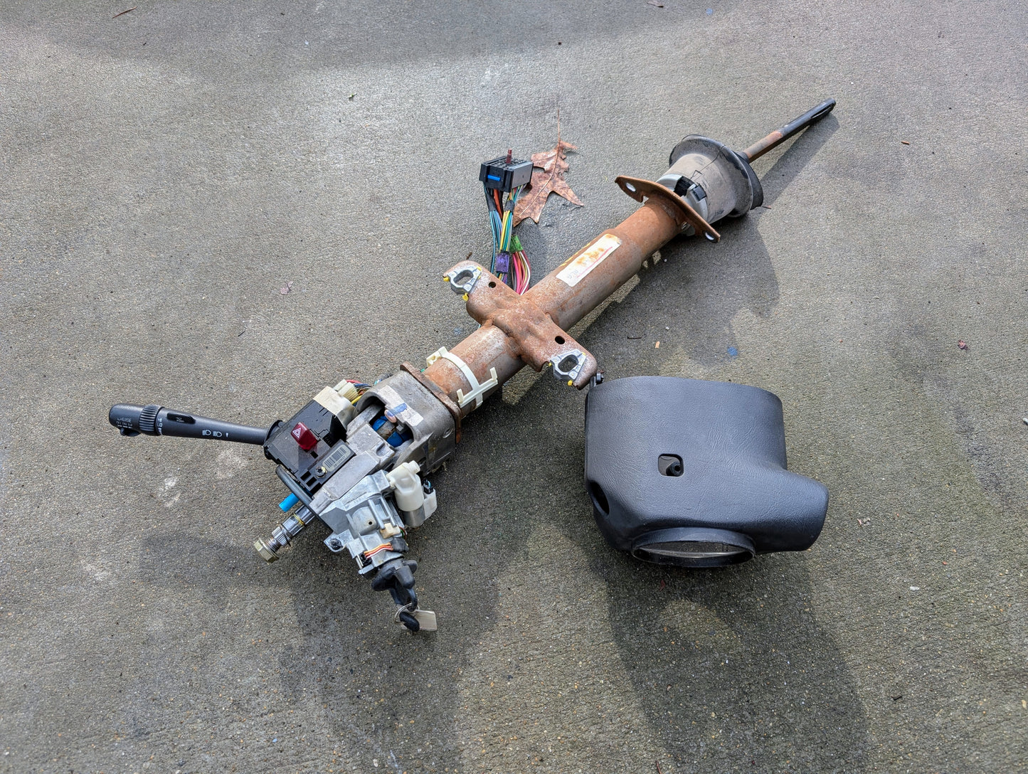 A General Motors OEM Steering Column w/ Tilt, Cruise Control, and Console Shift for 1998-2005 Chevy S10 Blazer, with attached wiring and turn signal lever. Detached black plastic cover included; item shown on concrete surface.