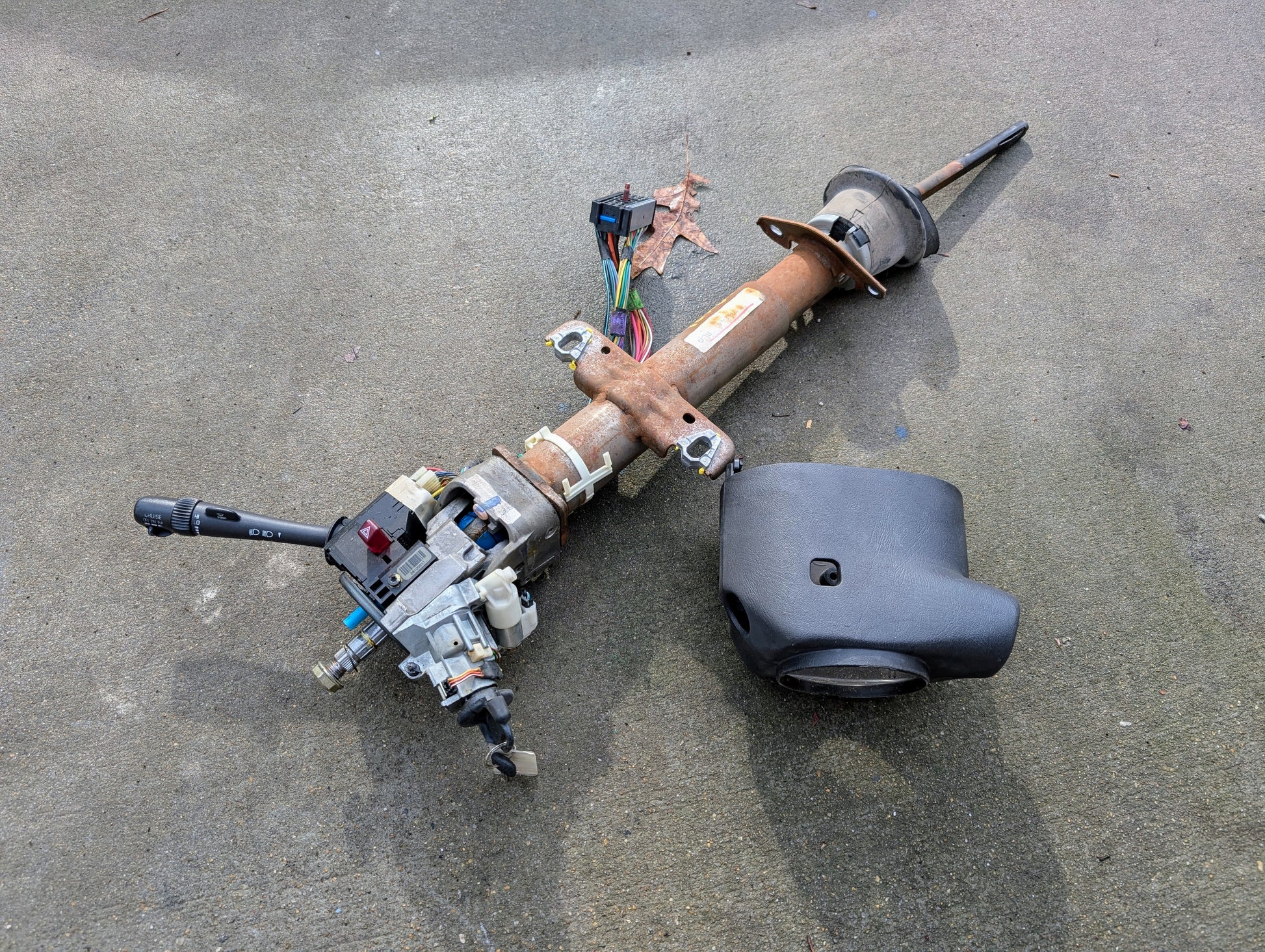 A General Motors OEM Steering Column w/ Tilt, Cruise Control, and Console Shift for 1998-2005 Chevy S10 Blazer, with attached wiring and turn signal lever. Detached black plastic cover included; item shown on concrete surface.