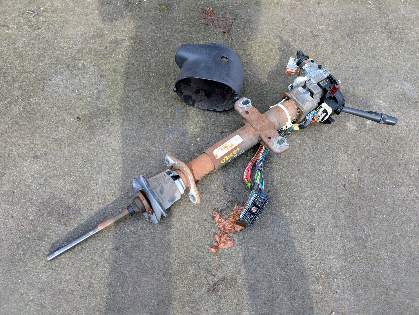 A disassembled General Motors OEM Steering Column w/Tilt, Cruise Control, and Console Shift for 1998-2005 Chevy S10 Blazer lies on concrete, with a black plastic cover and dry leaves nearby.