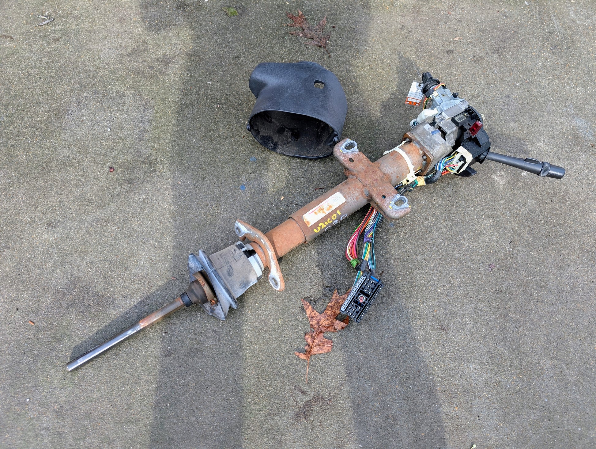 A disassembled General Motors OEM Steering Column w/Tilt, Cruise Control, and Console Shift for 1998-2005 Chevy S10 Blazer lies on concrete, with a black plastic cover and dry leaves nearby.