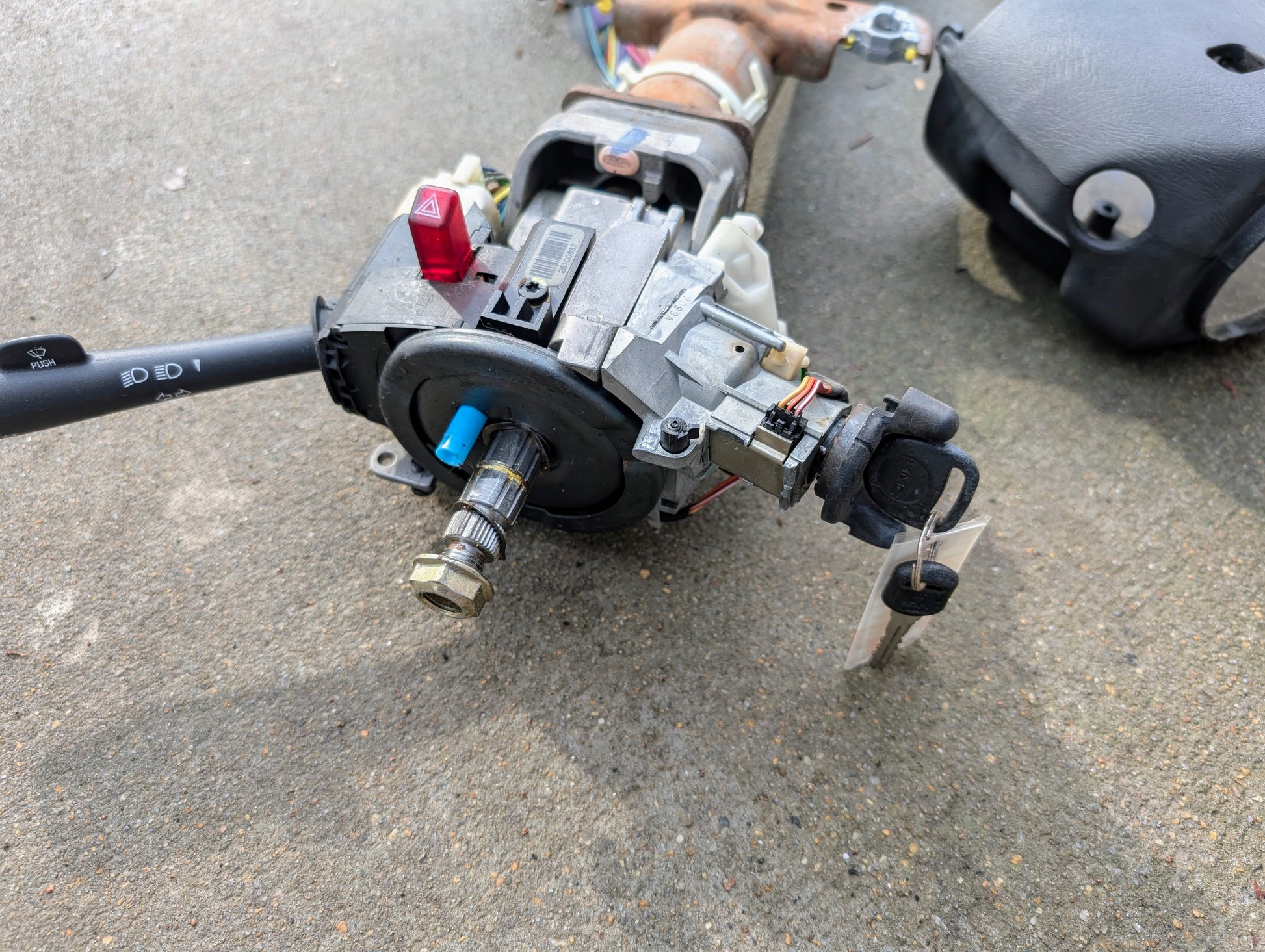 A General Motors OEM Steering Column with tilt, cruise control, and console shift for 1998-2005 Chevy S10 Blazer lies on concrete, along with an attached key, wiring, control stalk, and part of a black plastic cover.