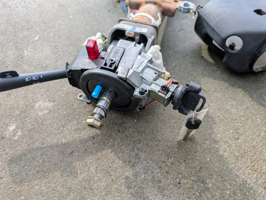 A General Motors OEM Steering Column with tilt, cruise control, and console shift for 1998-2005 Chevy S10 Blazer lies on concrete, along with an attached key, wiring, control stalk, and part of a black plastic cover.