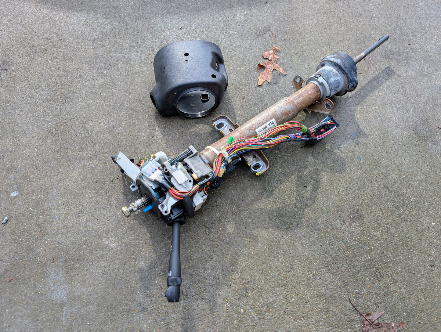 A disassembled General Motors OEM Steering Column w/Tilt, Cruise Control, and Console Shift for 1998-2005 Chevy S10 Blazer rests on concrete with exposed wiring, a black cover, and a brown leaf nearby.