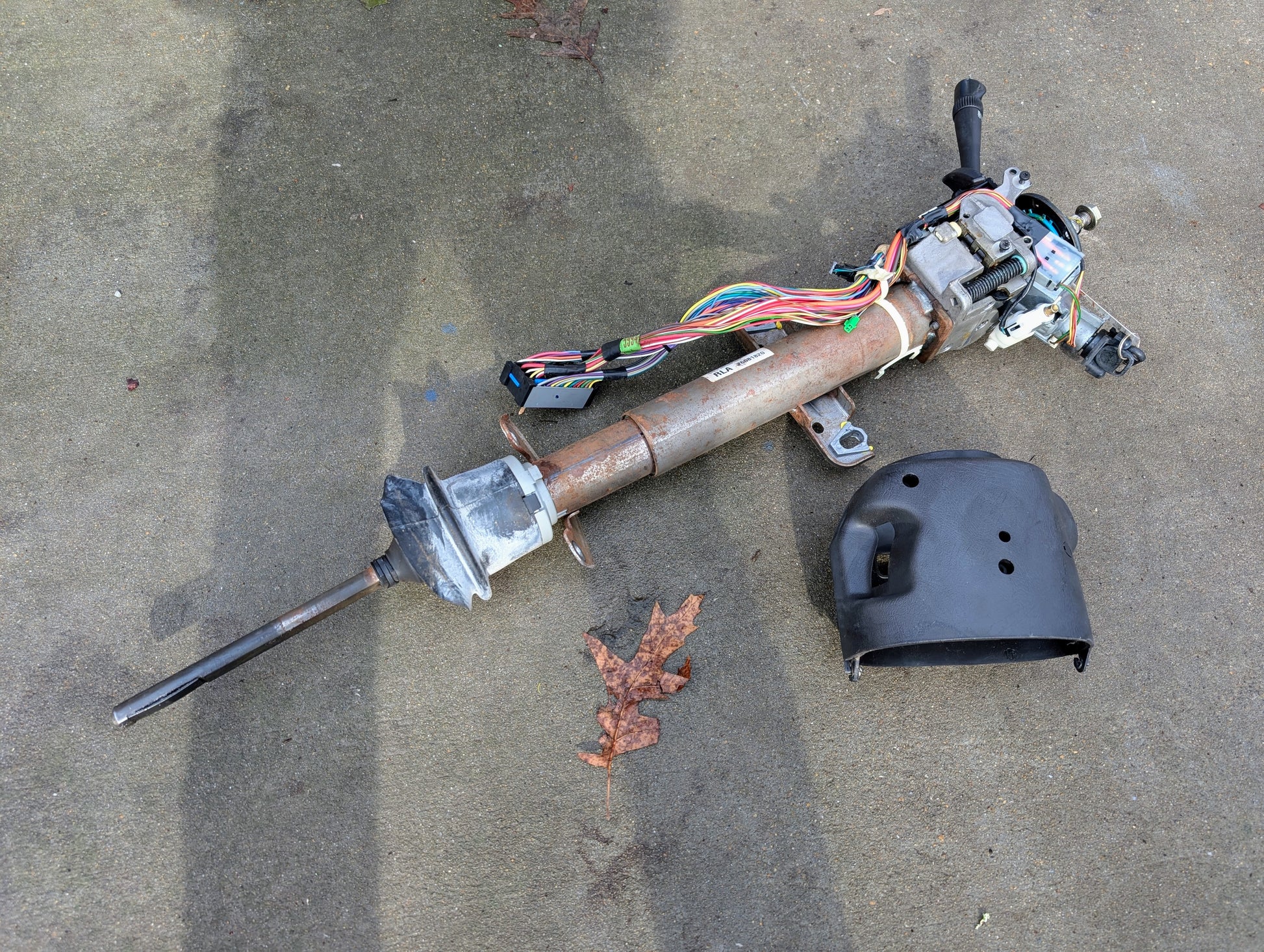A General Motors OEM Steering Column w/Tilt, Cruise Control, and Console Shift for 1998-2005 Chevy S10 Blazer and more, with wiring, rod, and cover, is disassembled on concrete near a dried leaf; fits automatic transmission models.
