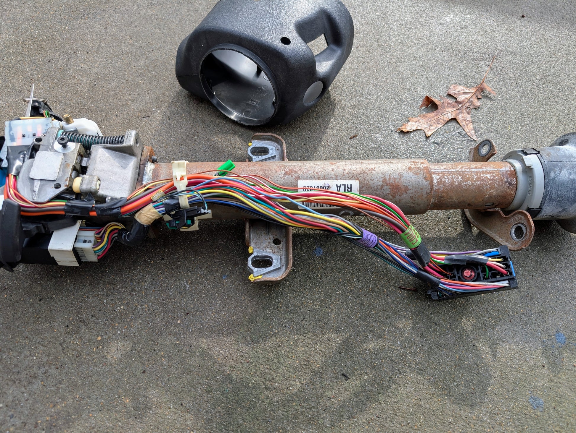 A General Motors OEM Steering Column w/Tilt, Cruise Control, and Console Shift for 1998-2005 Chevy S10 Blazer is partially disassembled on concrete with a plastic cover, wiring harness, connectors, and a dry leaf.