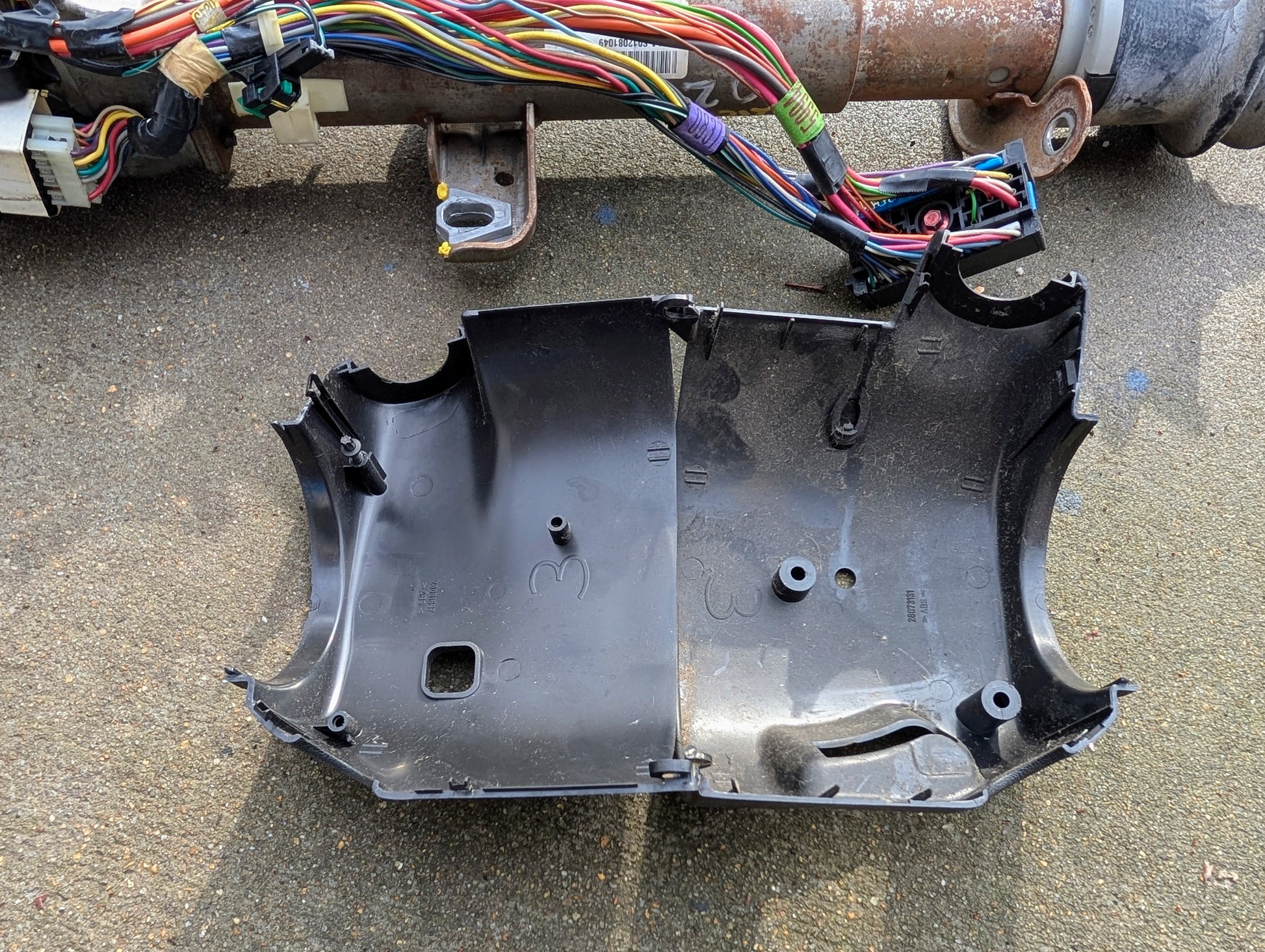 A black plastic steering column cover rests on rough concrete under a General Motors OEM Steering Column w/Tilt, Cruise Control, and Console Shift for 1998-2005 Chevy S10 Blazer. Colorful wiring and connectors are visible.