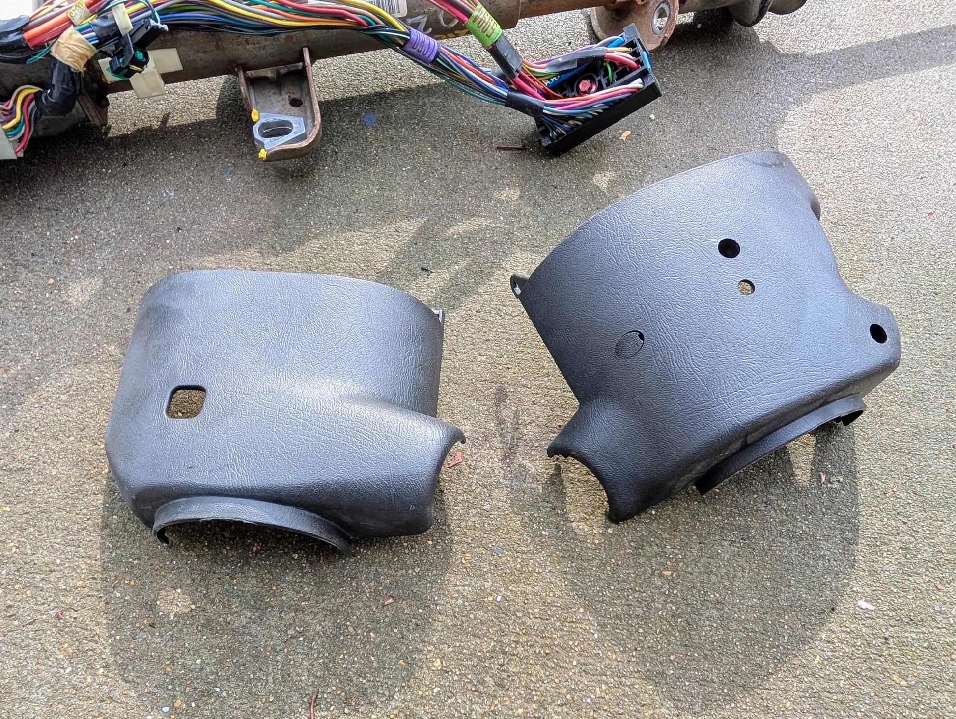 Two black plastic steering column covers from General Motors, made for the OEM Steering Column w/Tilt, Cruise Control & Console Shift (1998-2005 Chevy S10 Blazer), rest on concrete beside a colorful wiring harness.