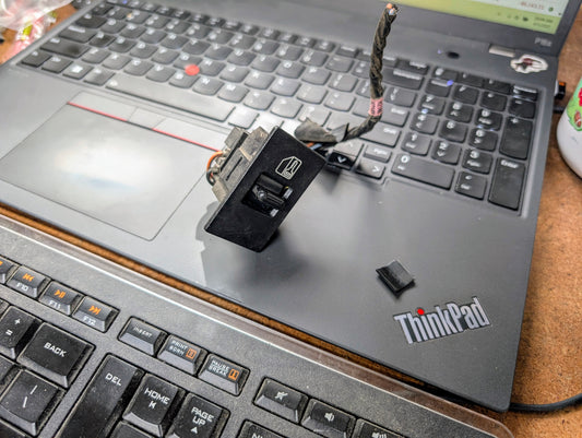 A General Motors OEM Driver LH Power Door Lock Switch for 1991-1994 Chevy S10 Blazer sits with its wire on the keyboard and touchpad of a Lenovo ThinkPad laptop, while a small loose plastic piece is nearby.