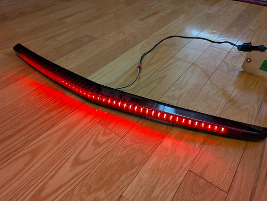 A General Motors OEM 2008-2013 Cadillac CTS Rear Stop Third Brake Light CHMSL is illuminated as rear lighting, placed on a wooden floor with its wiring visible.