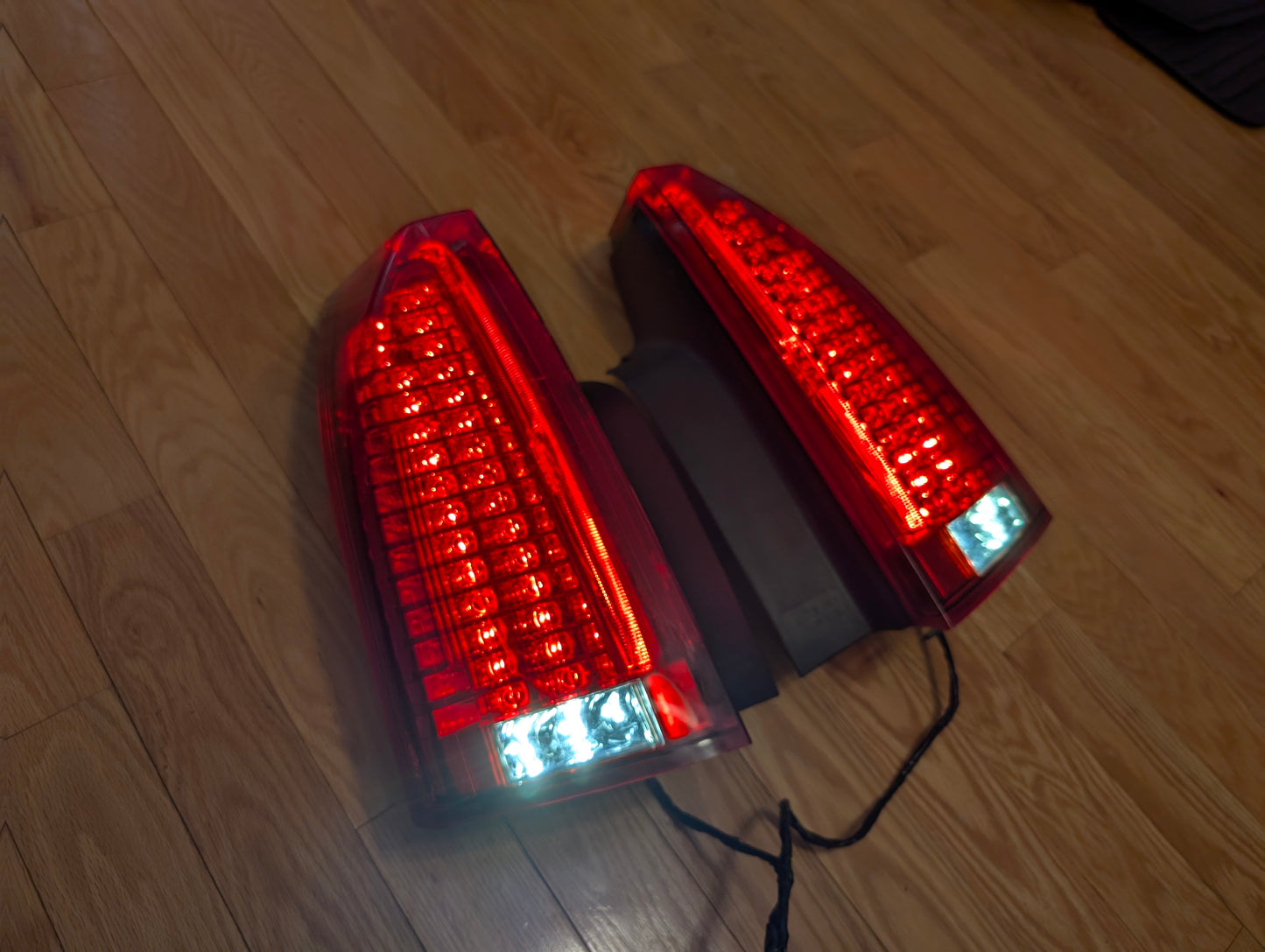 A pair of OEM left and right tail light brake lens assemblies by General Motors for 2008-2013 Cadillac CTS are shown with connected black wires, emitting a bright white-blue glow—ideal for rear lamp replacement or upgrade needs.