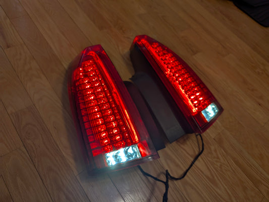 A pair of OEM left and right tail light brake lens assemblies by General Motors for 2008-2013 Cadillac CTS are shown with connected black wires, emitting a bright white-blue glow—ideal for rear lamp replacement or upgrade needs.