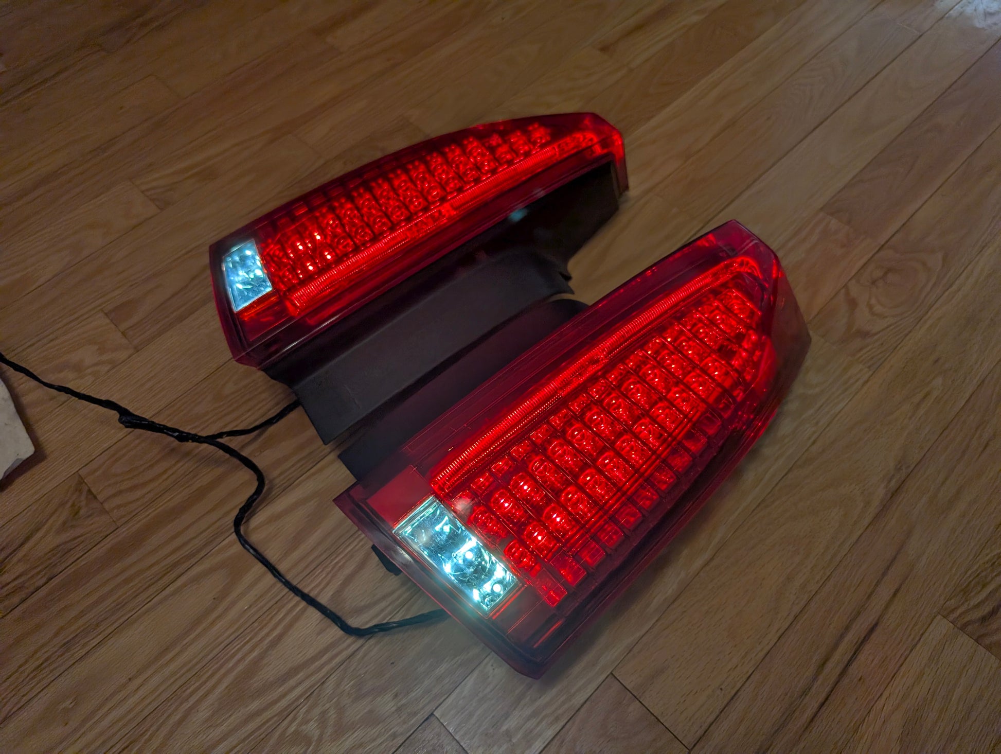 A pair of OEM General Motors left and right tail light brake lens assemblies for 2008-2013 Cadillac CTS are shown side by side on a wooden floor, illuminated with bright red and white LED lights and visible black wires.