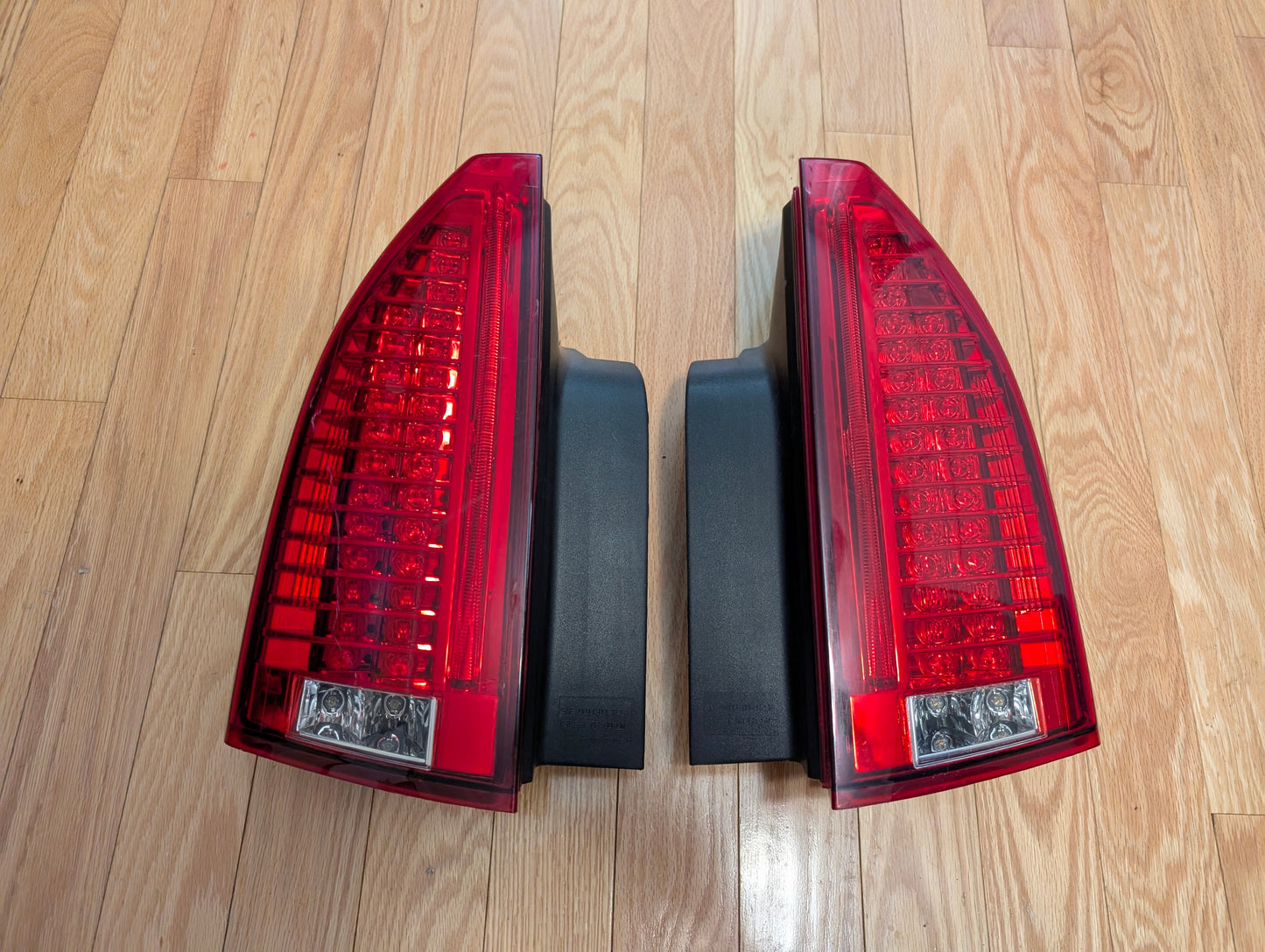 A pair of General Motors OEM left and right tail light brake lens assemblies for 2008-2013 Cadillac CTS are shown side by side on a light wood floor.