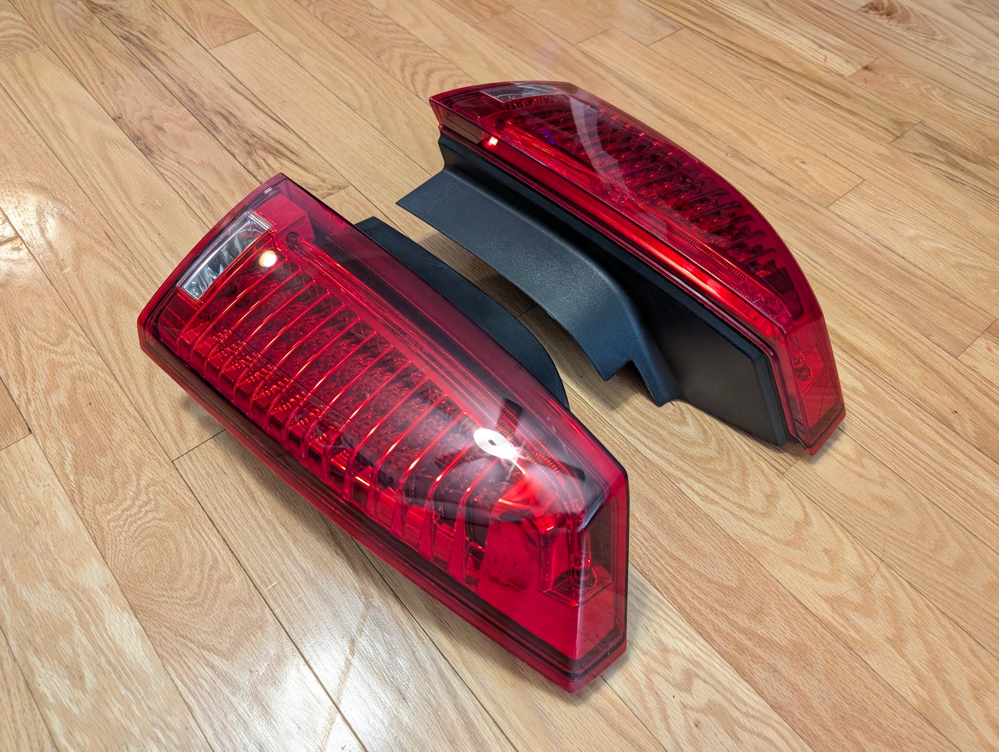 A pair of OEM General Motors left and right tail light brake lens assemblies for 2008-2013 Cadillac CTS are set side by side on a light wooden floor, each facing a different direction. Both lights have black backing.