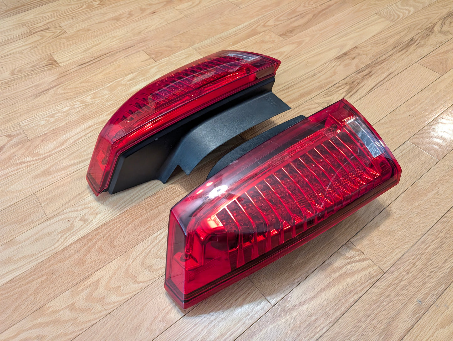 A pair of General Motors OEM left and right tail light brake lens assemblies for 2008–2013 Cadillac CTS are set parallel on a light wood floor, one slightly behind the other, ready for rear lamp replacement.