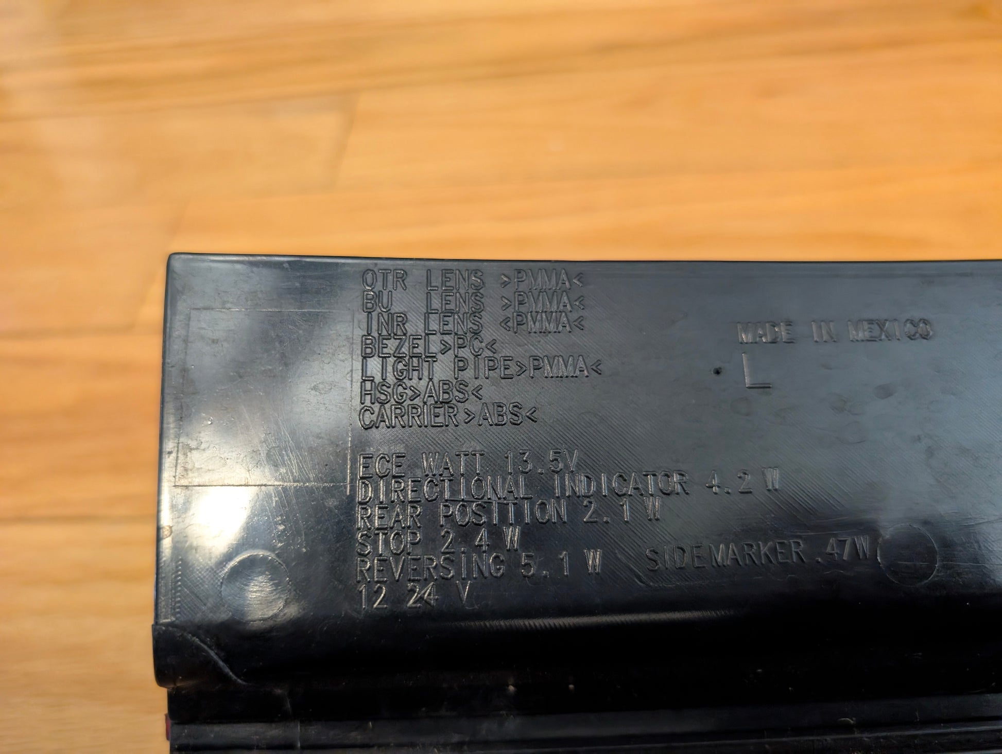 Close-up of a General Motors OEM pair left and right tail light brake lamp lens, tested for 2008-2013 Cadillac CTS; black plastic assembly with engraved specifications, shown on a wooden surface with some text hard to read due to lighting.