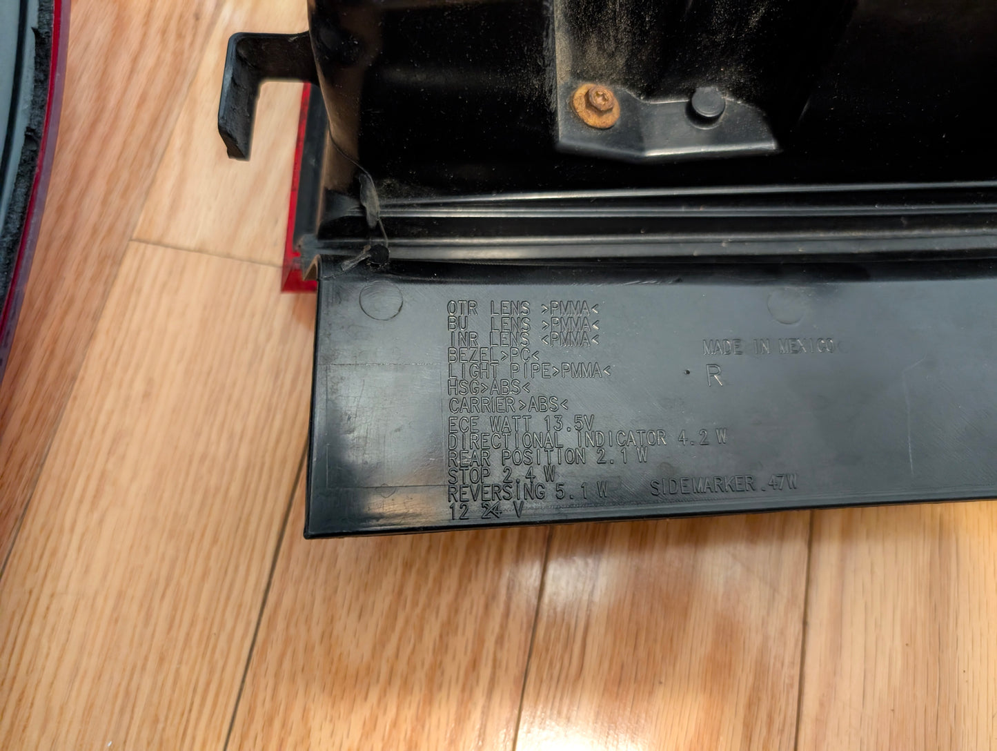 A close-up of a General Motors OEM left and right tail light lamp brake lens for 2008-2013 Cadillac CTS shows embossed specs and codes on the plastic part, with some dust on the assembly, placed on a wooden floor.