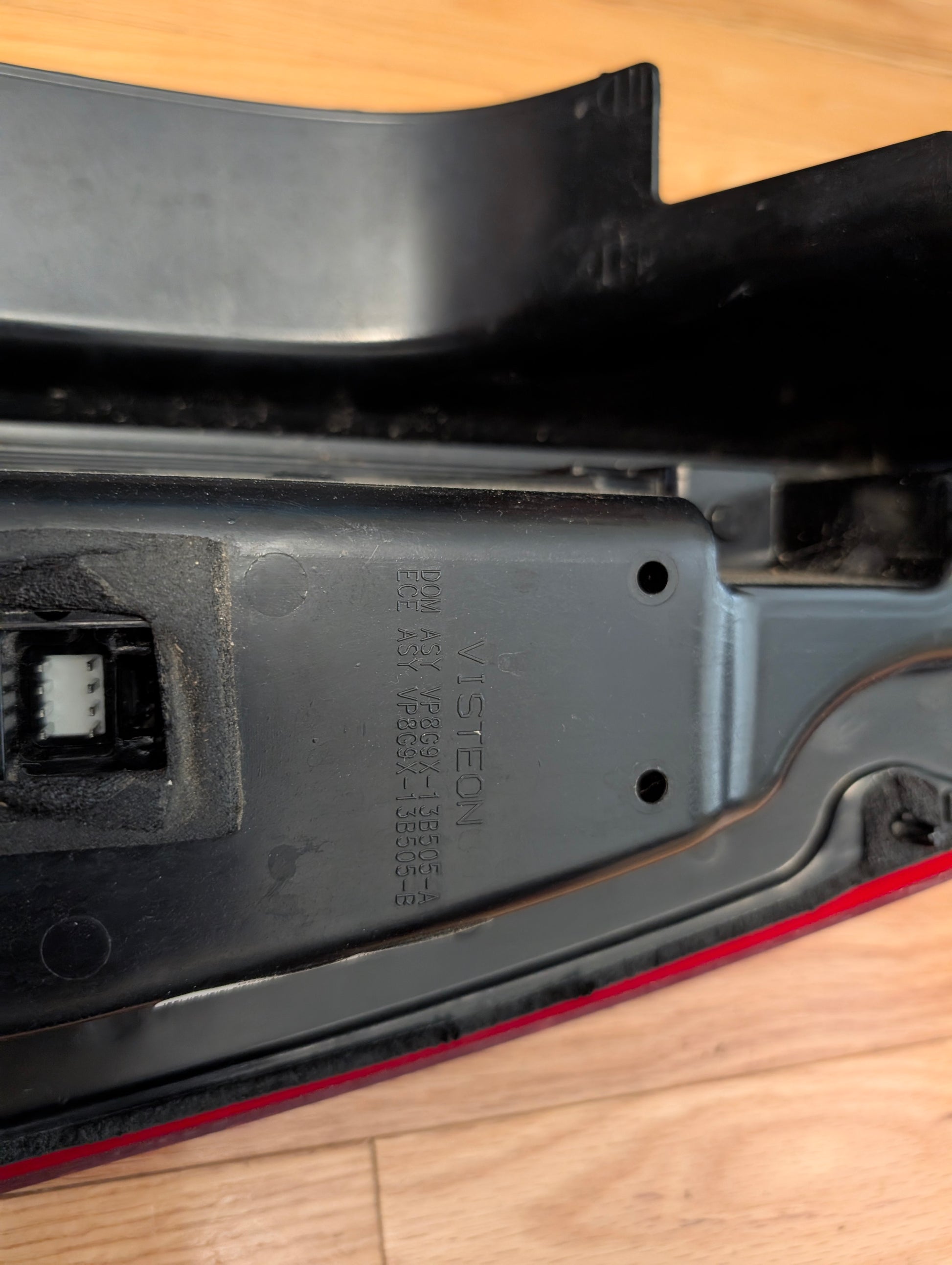 Close-up of a black plastic OEM Pair Left Right Tail Light Lamp Brake Lens by General Motors for 2008-2013 Cadillac CTS, with identification codes and VISTEON label molded on the surface; red edge rear lamp assembly visible on wooden floor.