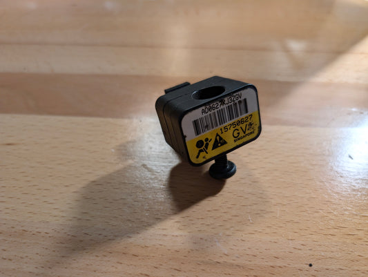 A small black General Motors OEM Front Impact Air Bag Sensor 15750627 for 2003-2004 Chevy Express/GMC Savanna, featuring a barcode label and yellow warning sticker, rests on a light wood surface with visible shadows.