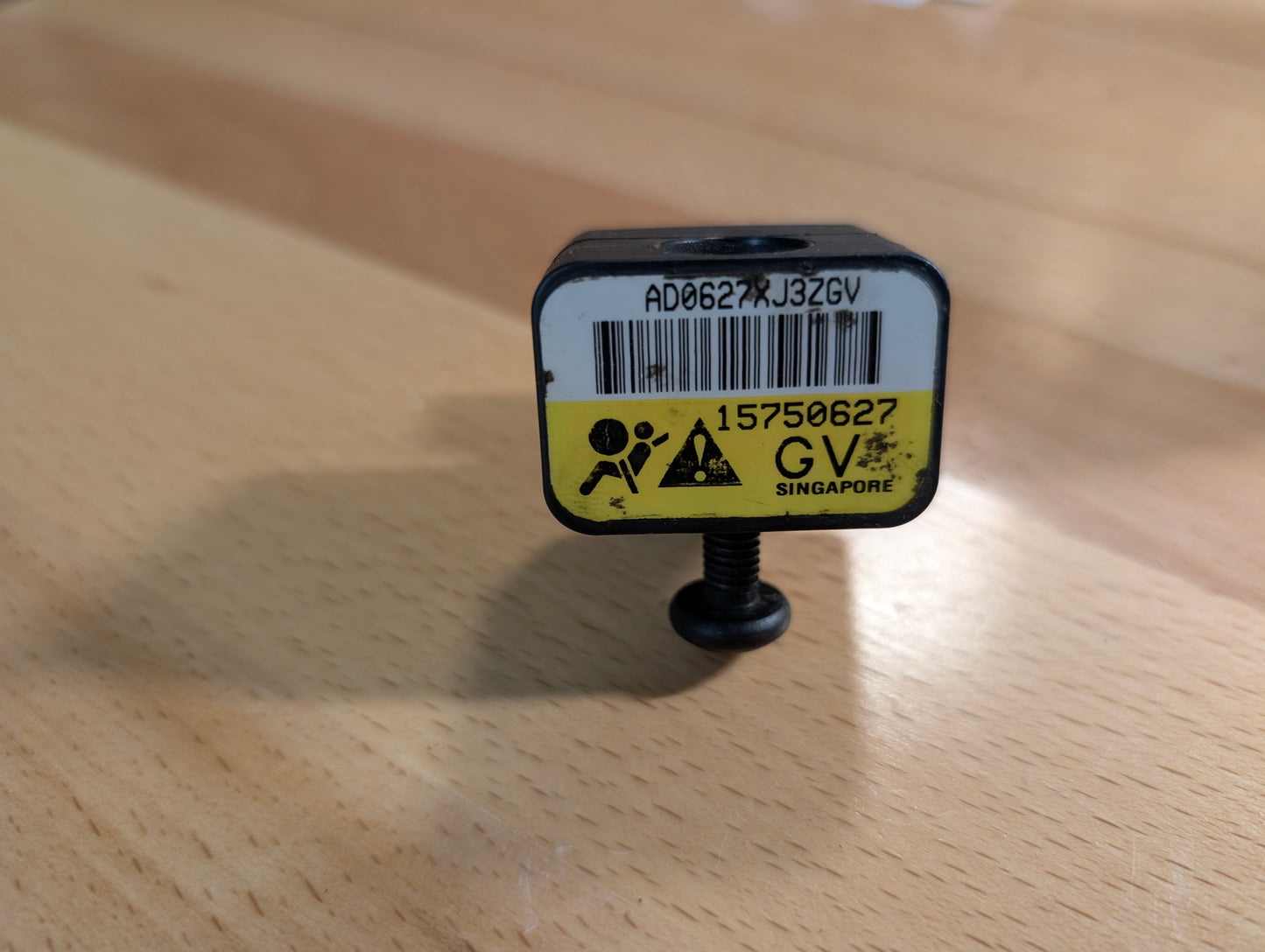 A small black General Motors OEM Front Impact Air Bag Sensor 15750627 for 2003-2004 Chevy Express or GMC Savanna, featuring a yellow label, barcode, bottom screw, and marked GV SINGAPORE, is shown on a wooden surface.