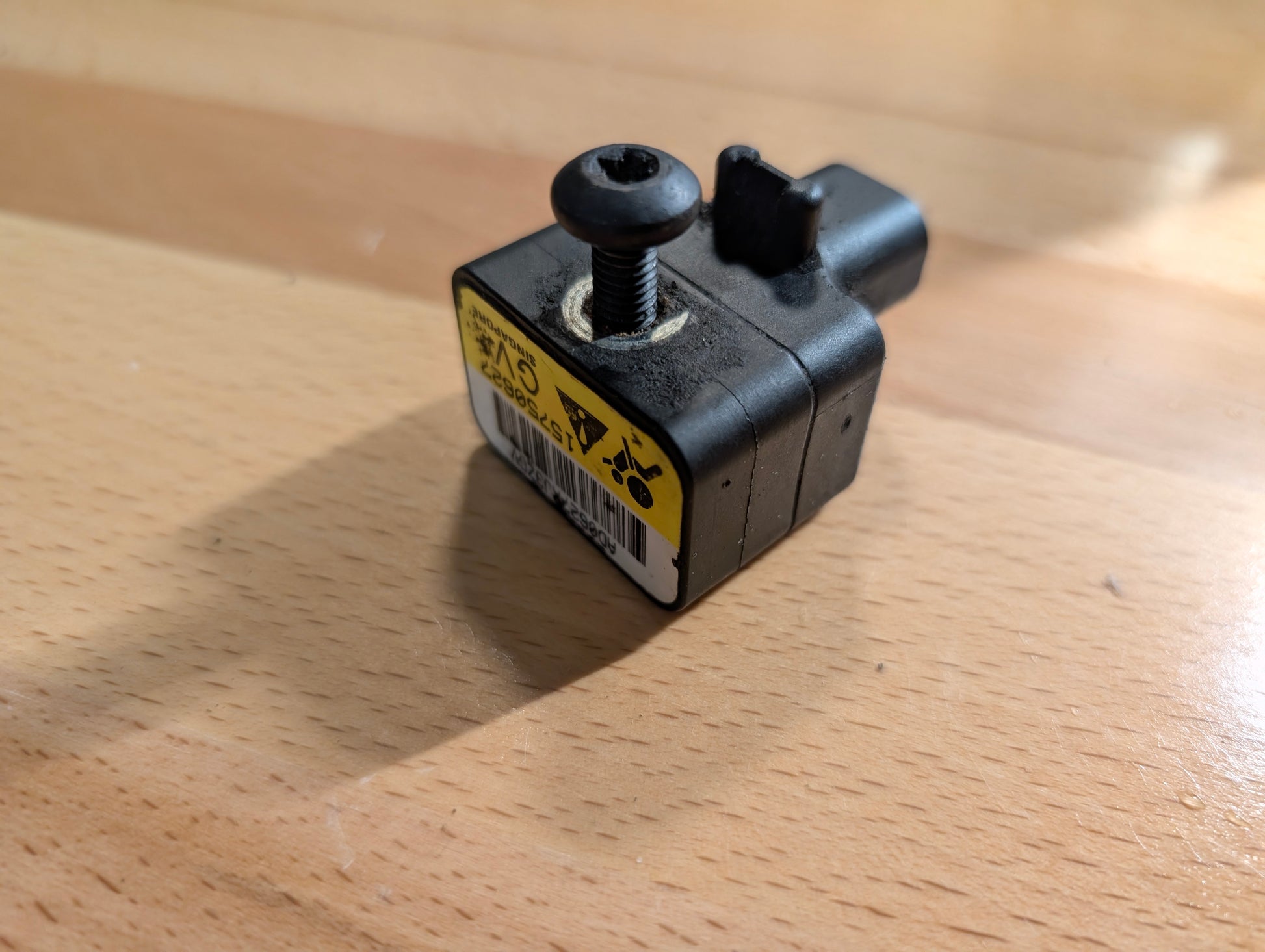 A compact black OEM Front Impact Air Bag Sensor 15750627 by General Motors for 2003-2004 Chevy Express/GMC Savanna, featuring a top screw and yellow caution label, sits on a light wooden surface with its shadow visible.