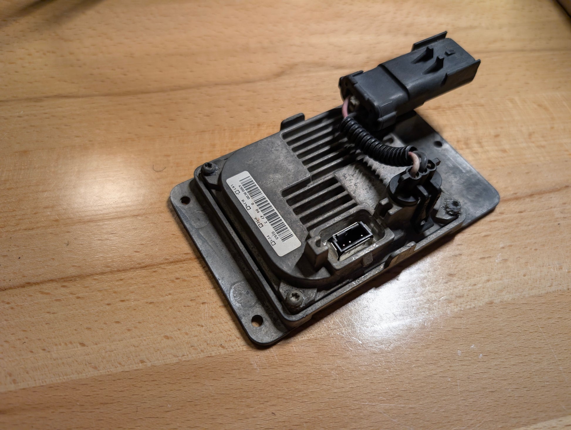 A close-up of a General Motors OEM HID Headlamp Ballast (part 19121438) for 2002-2006 Cadillac Escalade, showing its barcode label, connector port, and wiring on a light wooden surface—perfect for Escalade models.