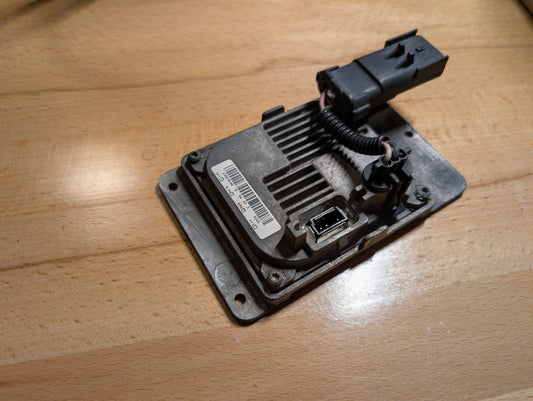 A close-up of a General Motors OEM HID Headlamp Ballast (part 19121438) for 2002-2006 Cadillac Escalade, showing its barcode label, connector port, and wiring on a light wooden surface—perfect for Escalade models.