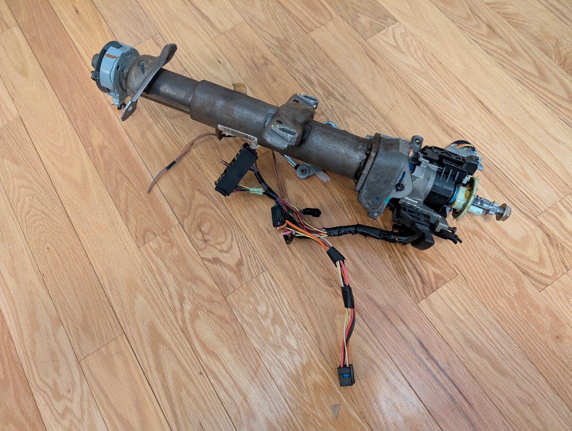An OEM Steering Column w/KEY for 2000-2002 GM Full Size Trucks by General Motors, featuring wiring harnesses, a column shifter, mounting brackets, and electrical connectors, is placed on a wood floor.