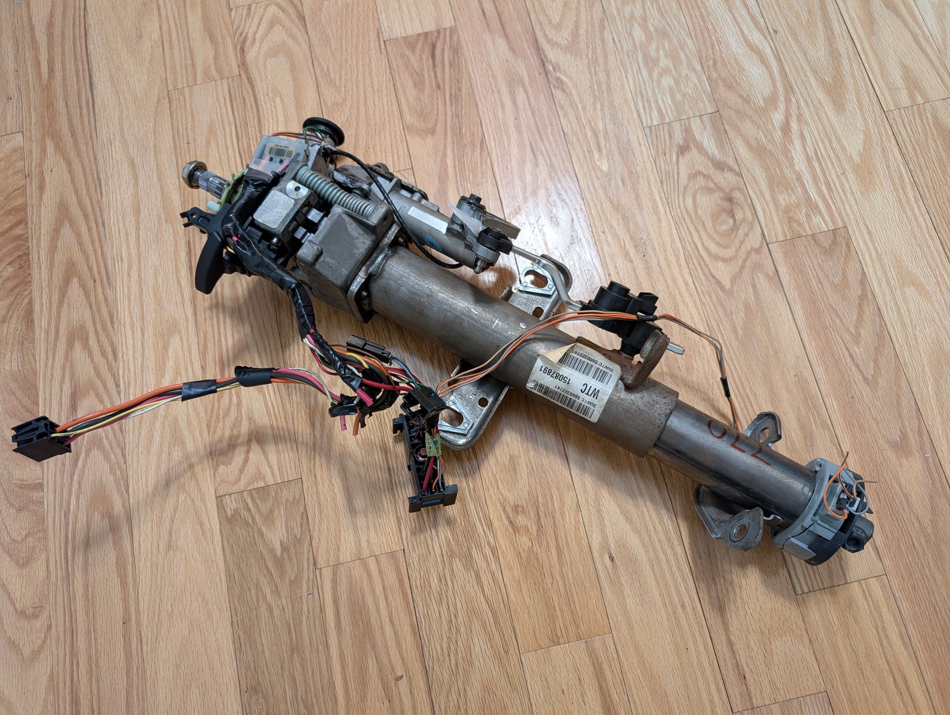 An OEM Steering Column w/KEY for 2000-2002 GM Full Size Trucks by General Motors, featuring a column shifter for automatic transmission, wiring, electronic components, mounting brackets, and connectors rests on a light hardwood floor.