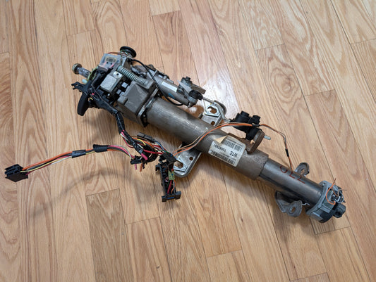 An OEM Steering Column w/KEY for 2000-2002 GM Full Size Trucks by General Motors, featuring a column shifter for automatic transmission, wiring, electronic components, mounting brackets, and connectors rests on a light hardwood floor.