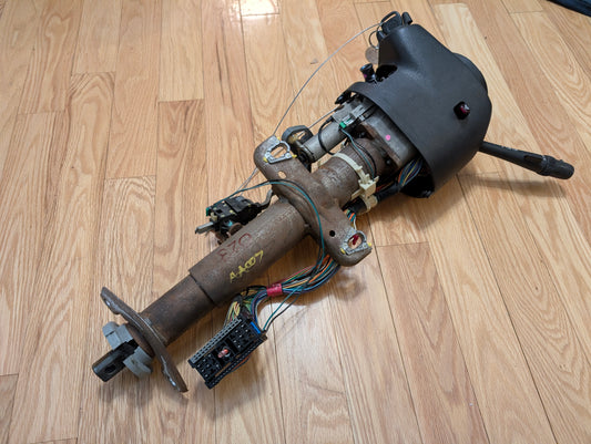 A General Motors OEM Steering Column w/Tilt, Cruise Control, and Column Shift for 1998-2005 Chevy S10 Blazer and more, including switches and black plastic housing, is shown lying on a wooden floor.