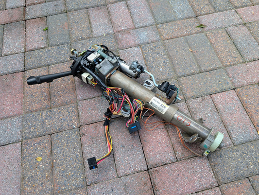 A General Motors OEM Steering Column for 2000-2002 GM Full Size Trucks, featuring switches and wiring harness for automatic transmission, is displayed on a brick surface, showing colored wires, connectors, and mechanical parts.