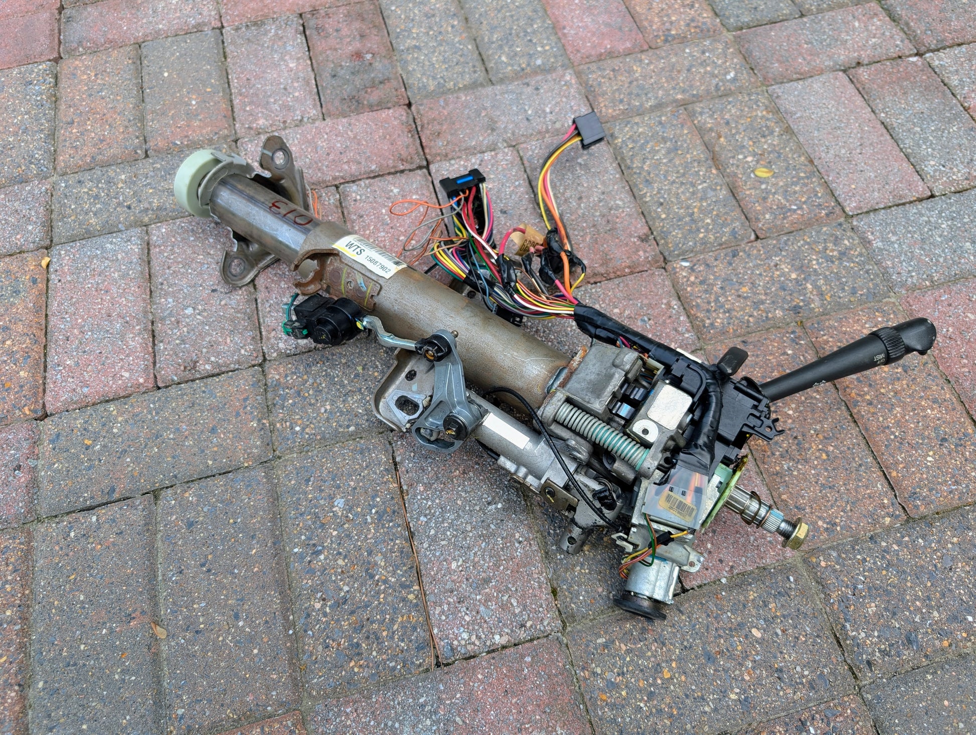 A OEM Steering Column for 2000-2002 GM Full Size Trucks by General Motors, complete with attached wiring and connectors, is shown disassembled on red and gray brick pavement after removal from an automatic transmission vehicle.