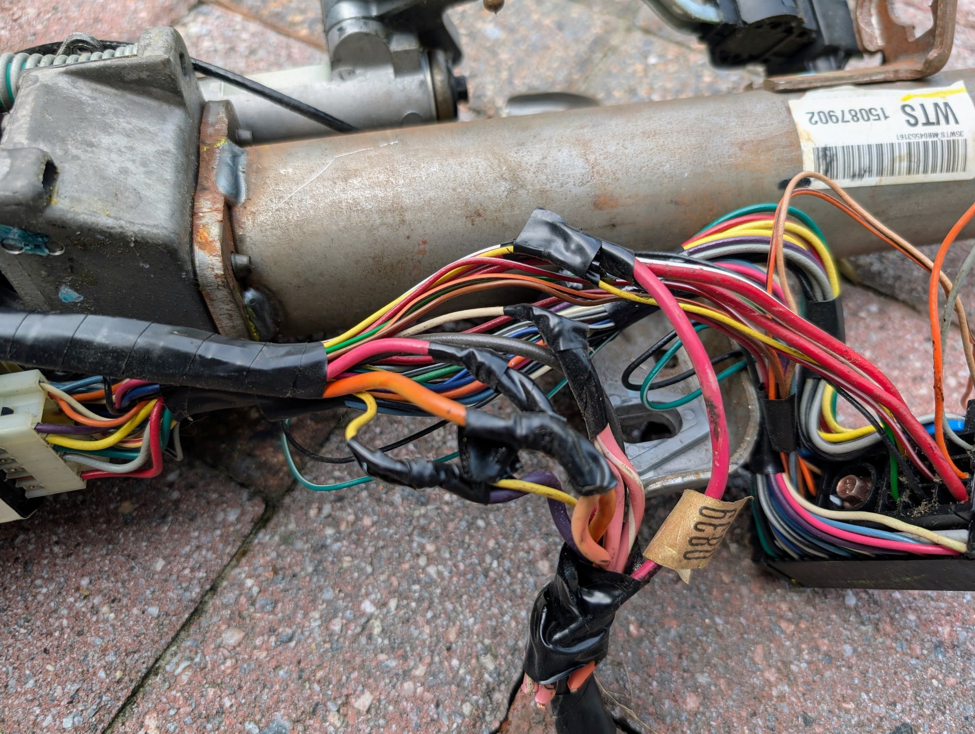 Close-up of a bundle of multicolored wires wrapped in black tape, attached to an OEM Steering Column for 2000-2002 GM Full Size Trucks by General Motors, resting on a textured brick surface.