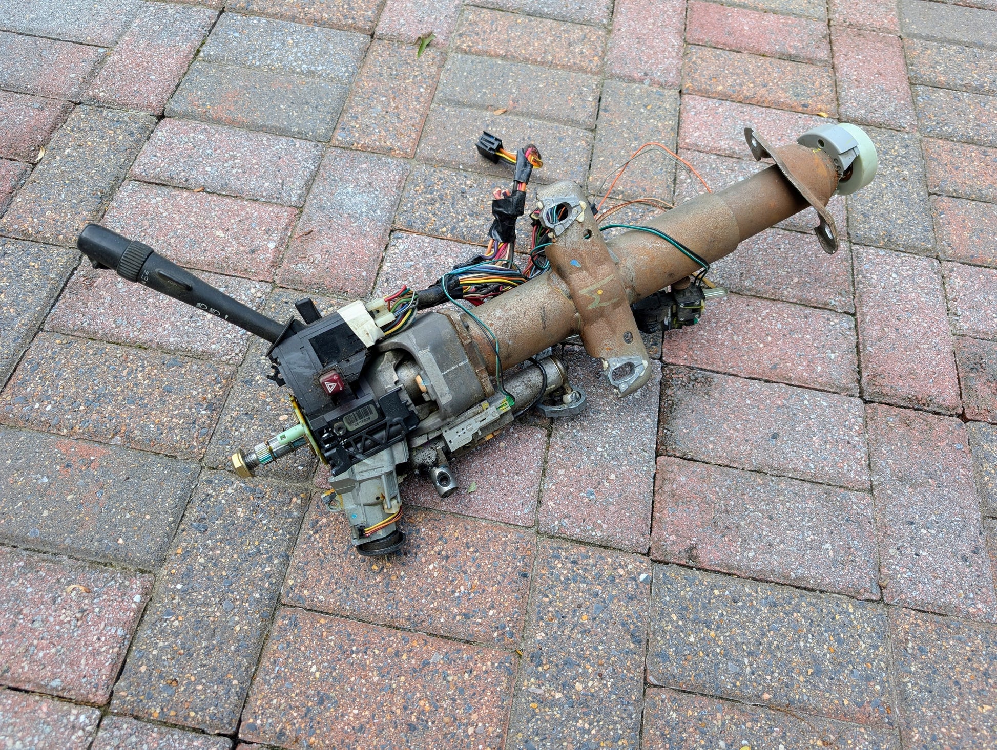 A General Motors OEM Steering Column for 2000-2002 GM Full Size Trucks with wiring and levers rests on a multicolored brick pavement.