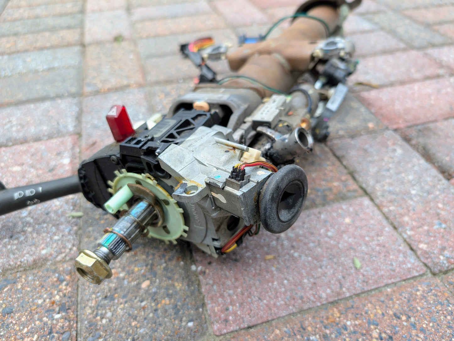 A close-up of a disassembled General Motors OEM Steering Column for 2000-2002 GM Full Size Trucks, showing wiring, gears, and metal parts on a brick-paved outdoor surface.