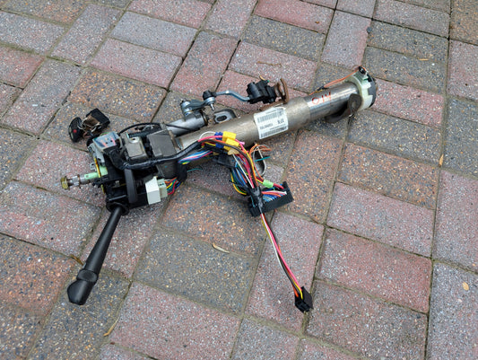An OEM Steering Column w/KEY for 2000-2002 GM Full Size Trucks by General Motors, with wiring, connectors, and control modules attached, is shown disassembled on brick pavement with automatic transmission components visible.