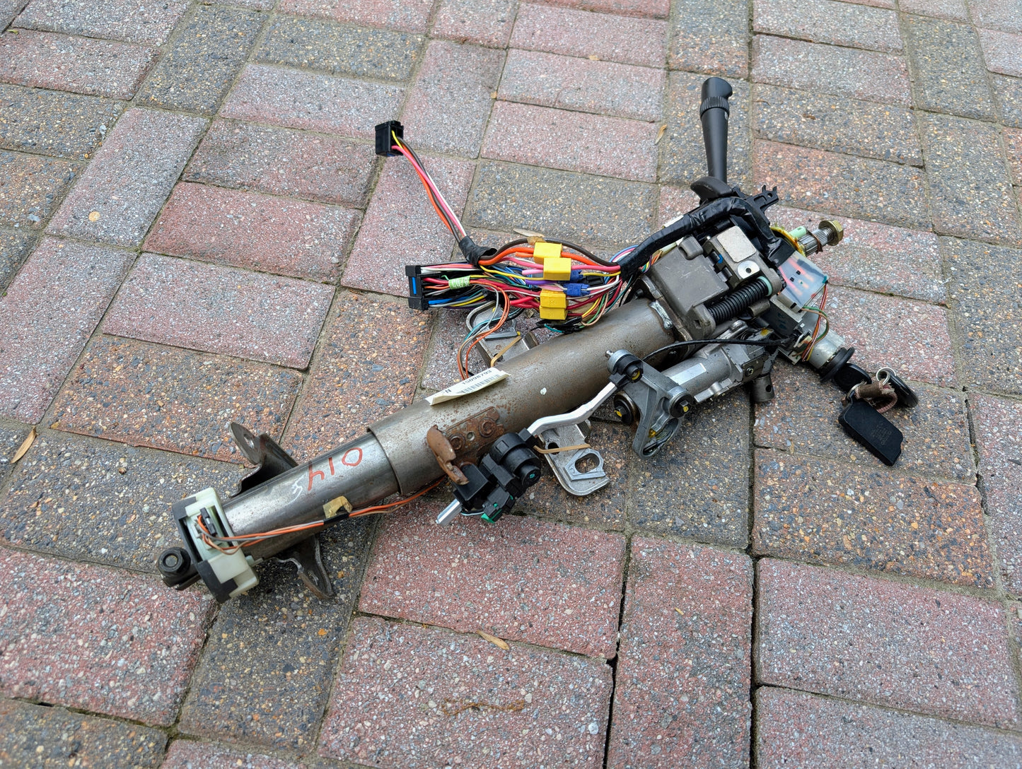 A General Motors OEM Steering Column w/KEY for 2000-2002 GM Full Size Trucks is disassembled with wiring and ignition switch, lying on a brick-paved surface.
