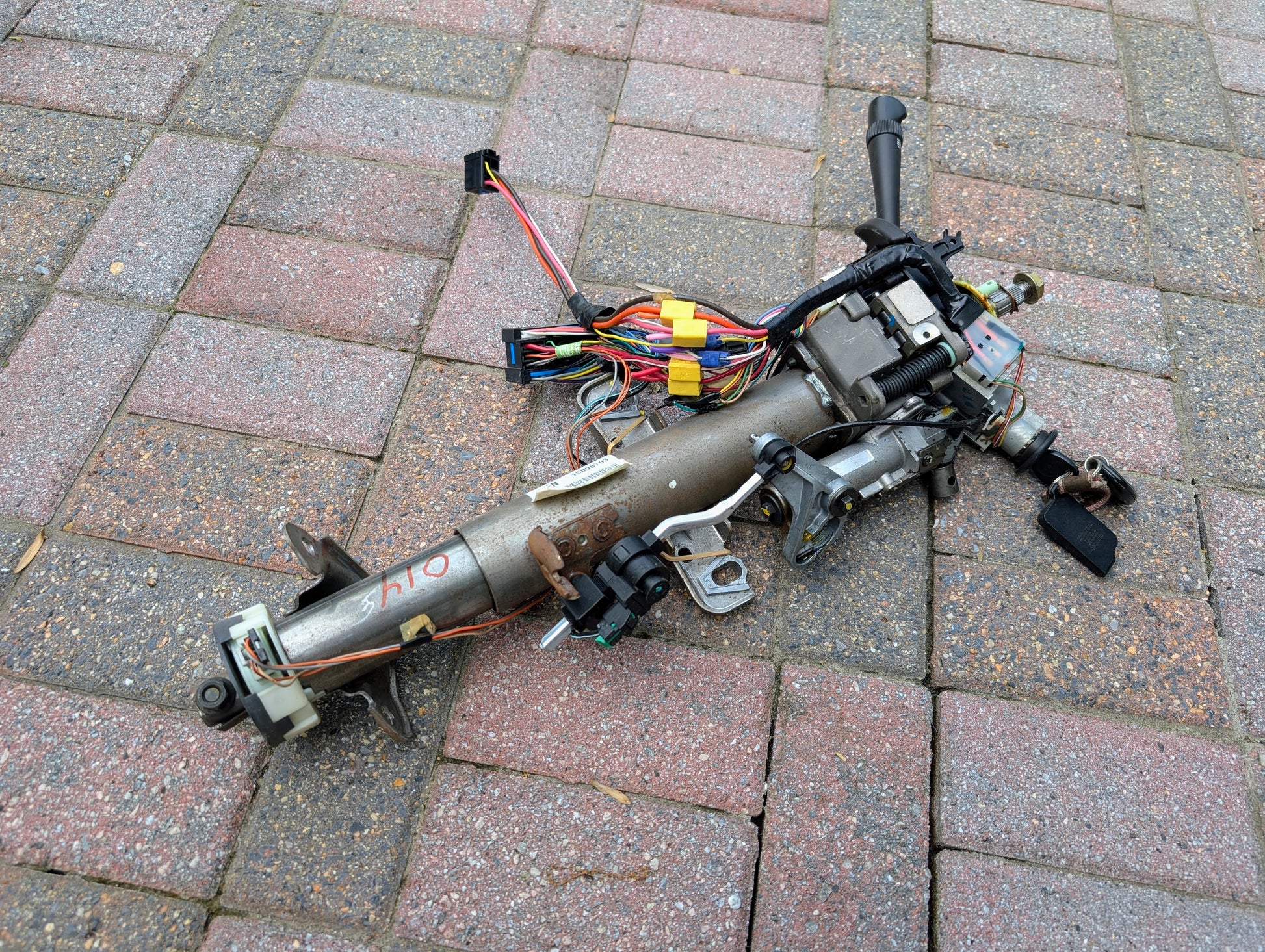 A General Motors OEM Steering Column w/KEY for 2000-2002 GM Full Size Trucks is disassembled with wiring and ignition switch, lying on a brick-paved surface.